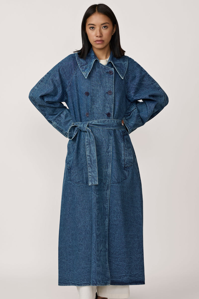 A person standing wearing a double-breasted, wide collar, A-line silhouette trench coat in washed denim blue.