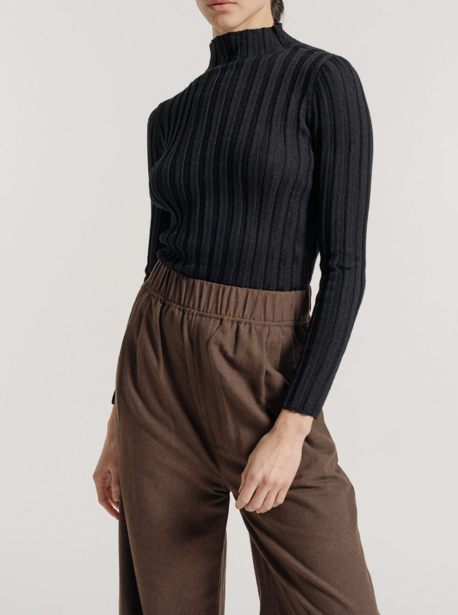 The model is wearing a vintage-inspired classic black Soa Ribbed Turtleneck top and brown wide leg pants made from ethically-sourced baby alpaca.