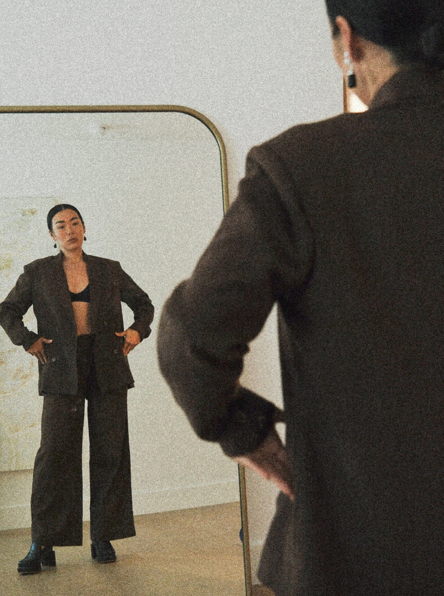 A woman is dressed in a Bea Blazer - Basalt Brown suit, admiring her tailored waistline in front of a mirror.