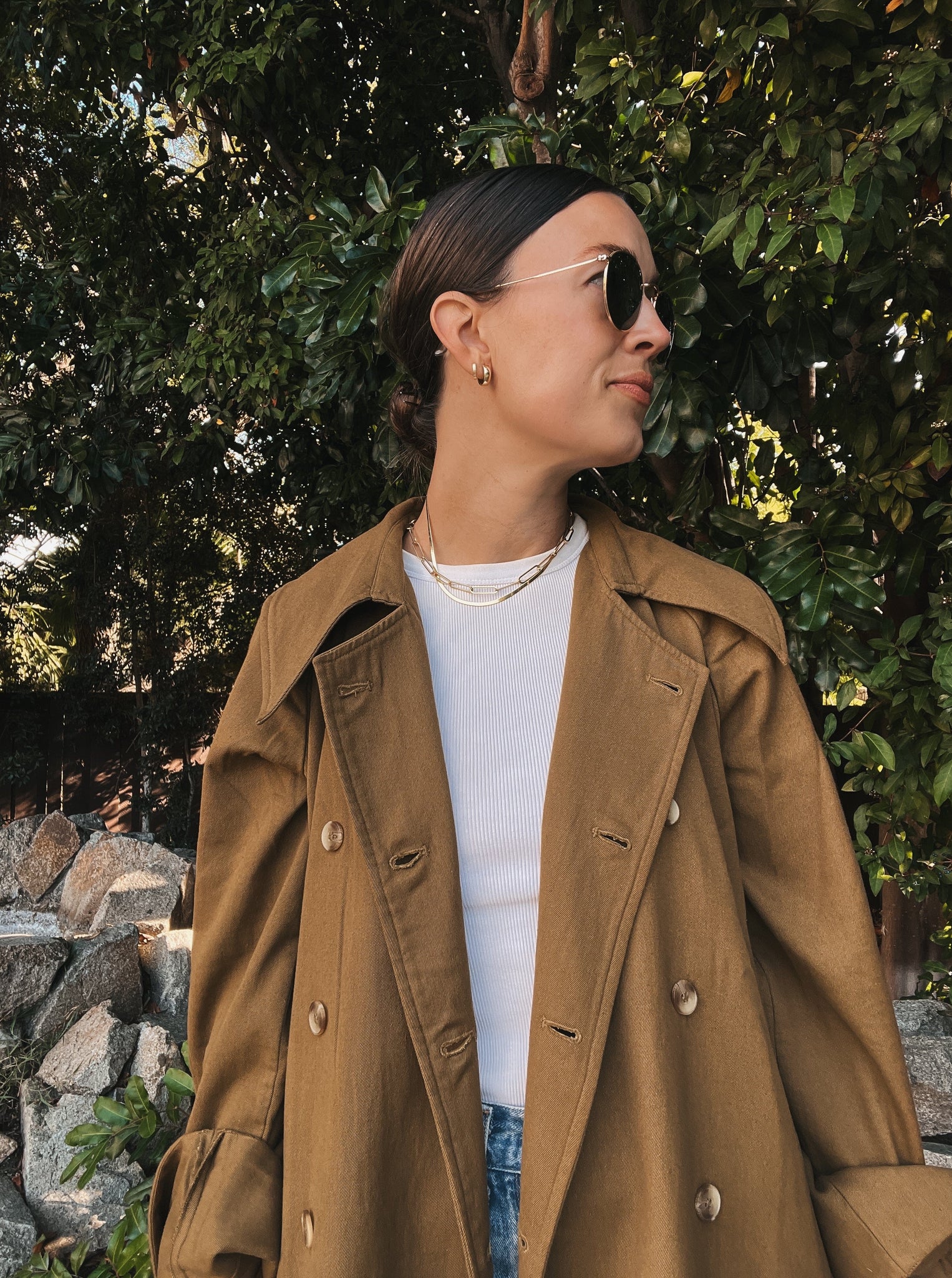 A woman wearing a Marin Trench Coat - Olive made in India tan trench coat made of cotton twill and matching jeans.