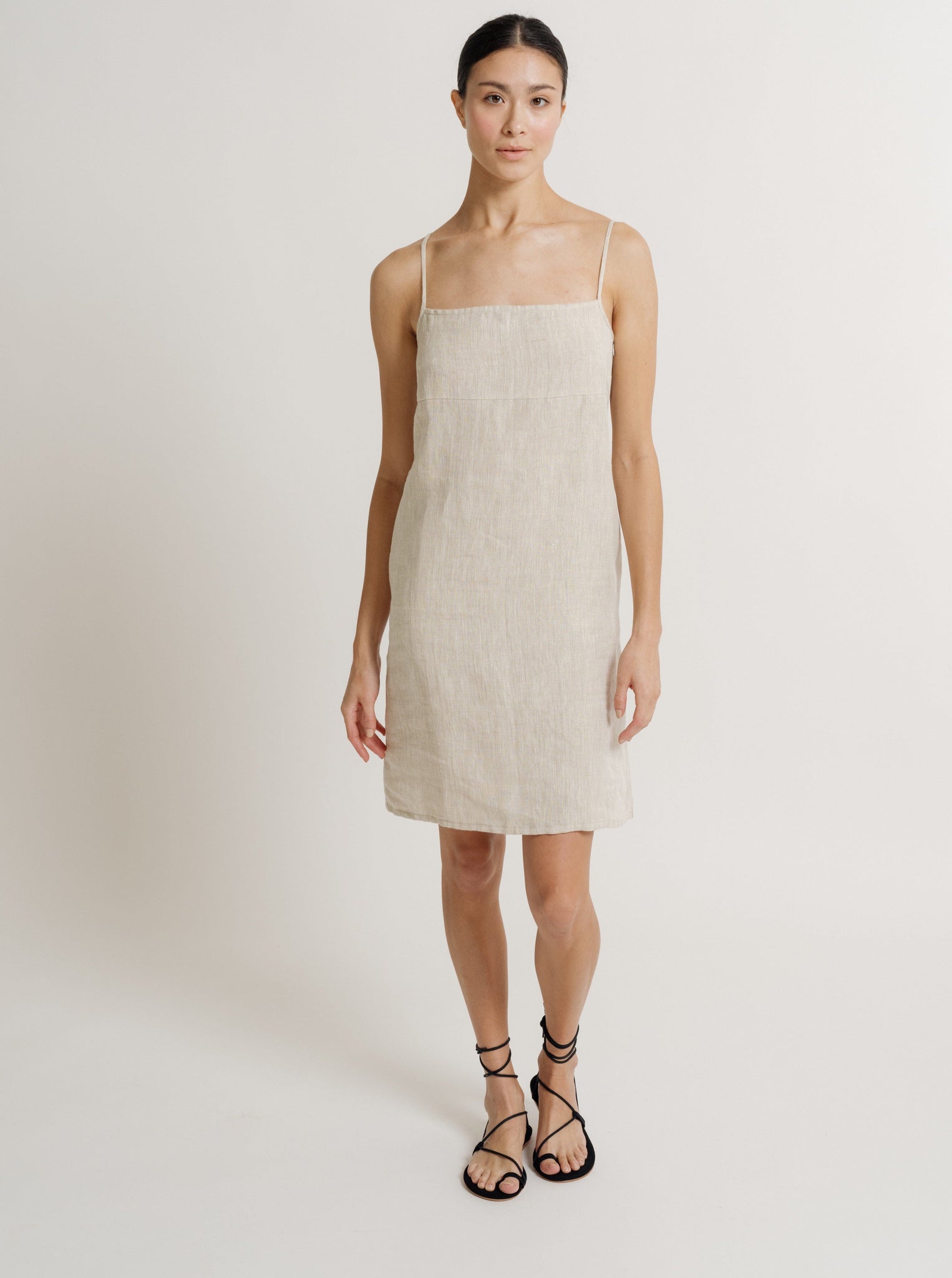 Woman in a like-new, Linen Mini Dress - Natural - Sample and black sandals standing against a neutral background for a photoshoot.