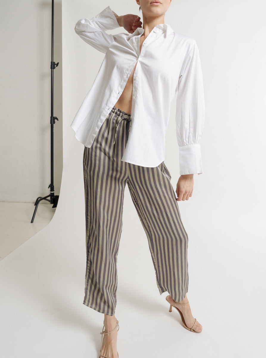 The model is wearing a white Museo Button Up - White Poplin shirt, made from organic cotton, paired with striped pants. The shirt has a tunic length.