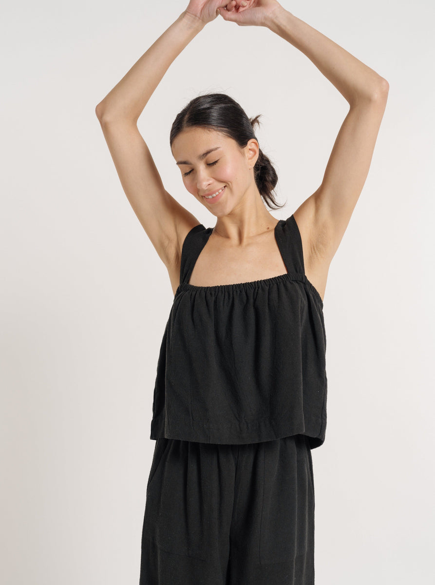A woman wearing a Black Silk Noil jumpsuit made of Cross Back Tank - Black Silk Noil - Pre-order, available for resort pre-orders and featuring a Cross Back Tank design.