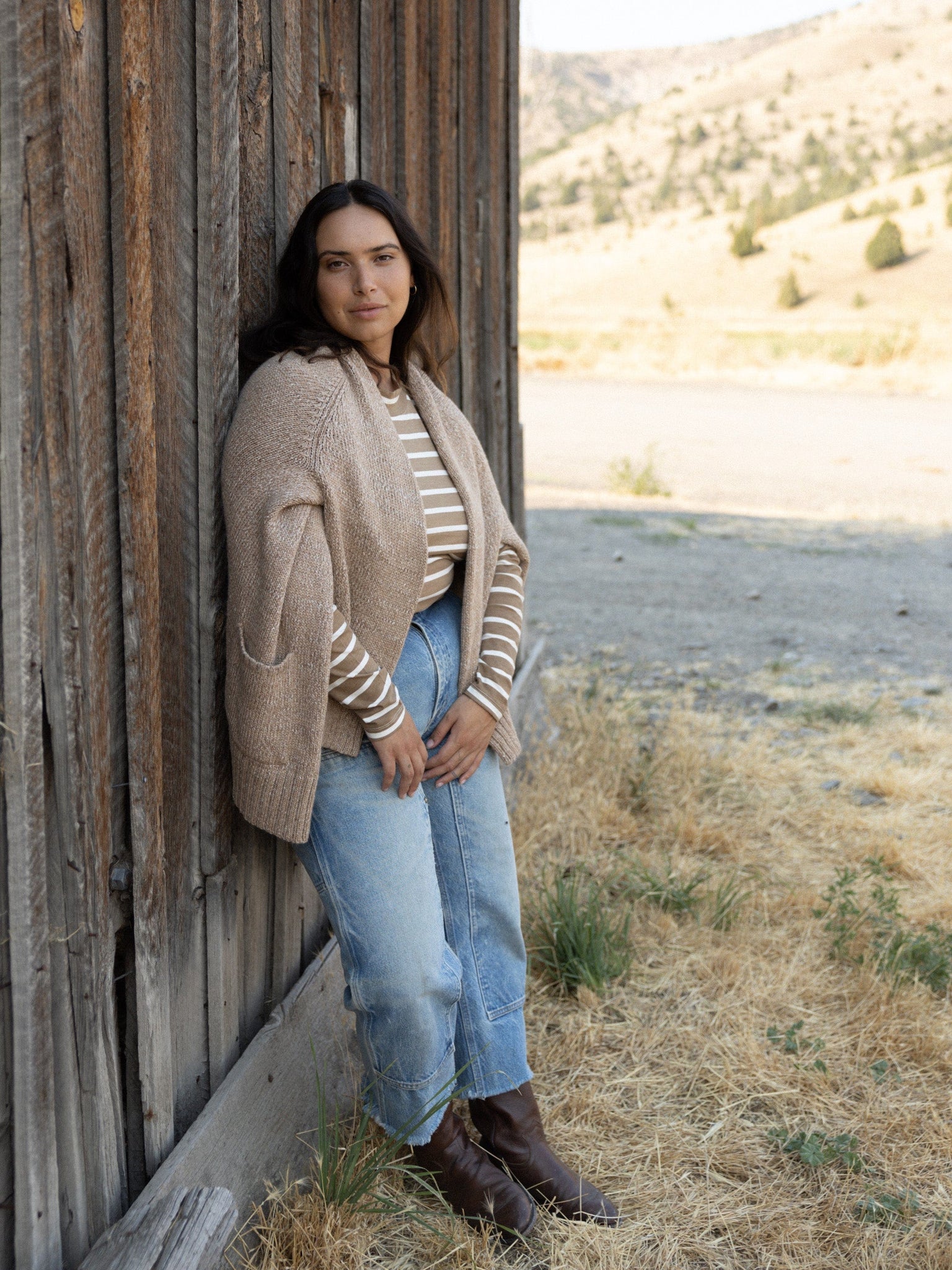 A fit woman wearing jeans and a cardigan leaning against a wooden wall while donning the Sample 144 - Mock Neck Long Sleeve Tee - Tannin Stripe.
