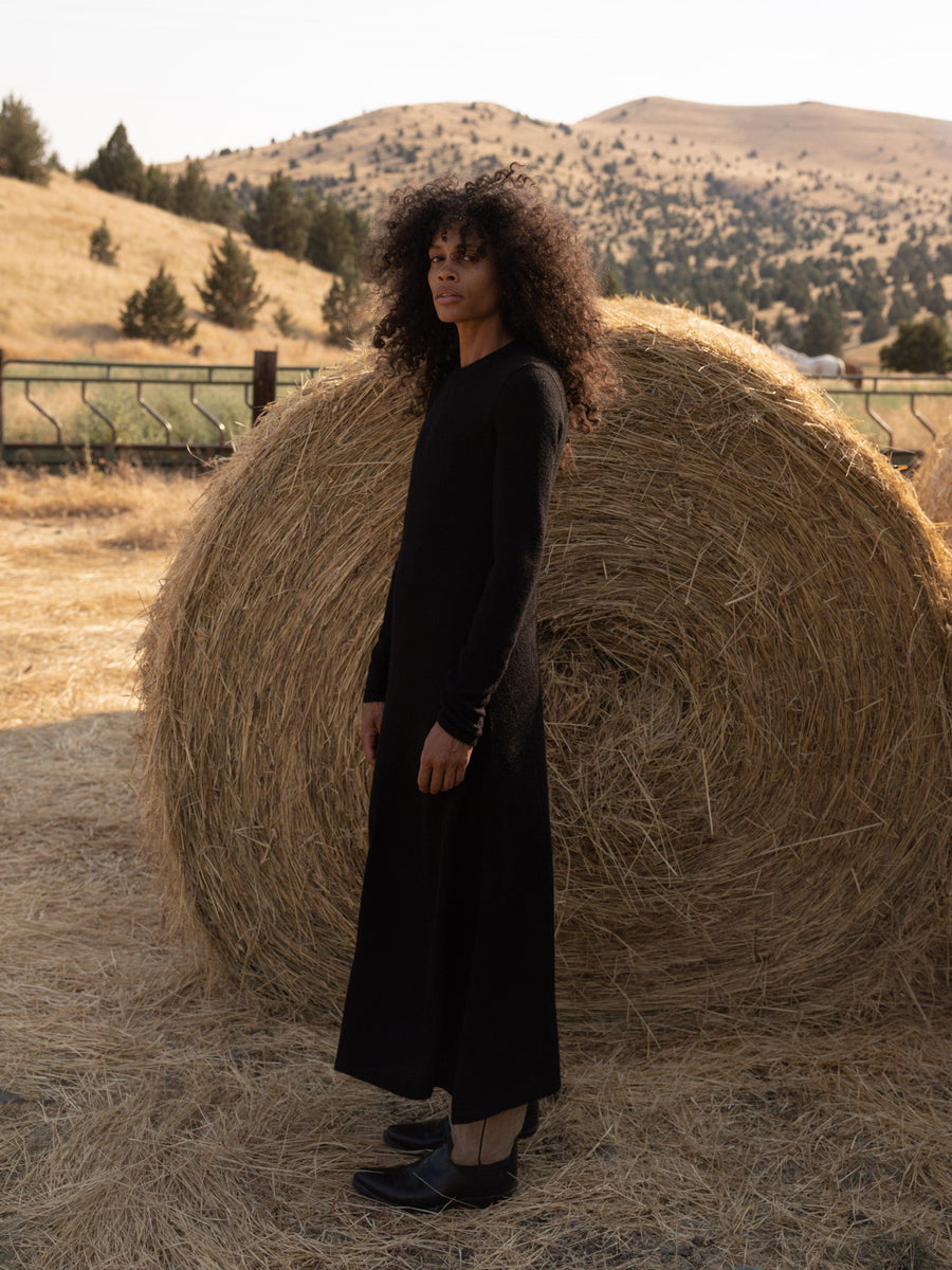 A woman in an ethically-sourced Ennis Dress - Black, standing next to a bale of hay.
