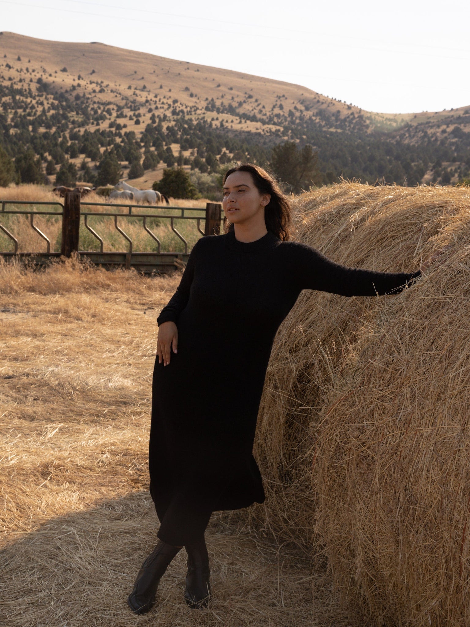 A woman ethically-sourced posing in front of hay bales wearing the Ennis Dress - Black made of baby alpaca.