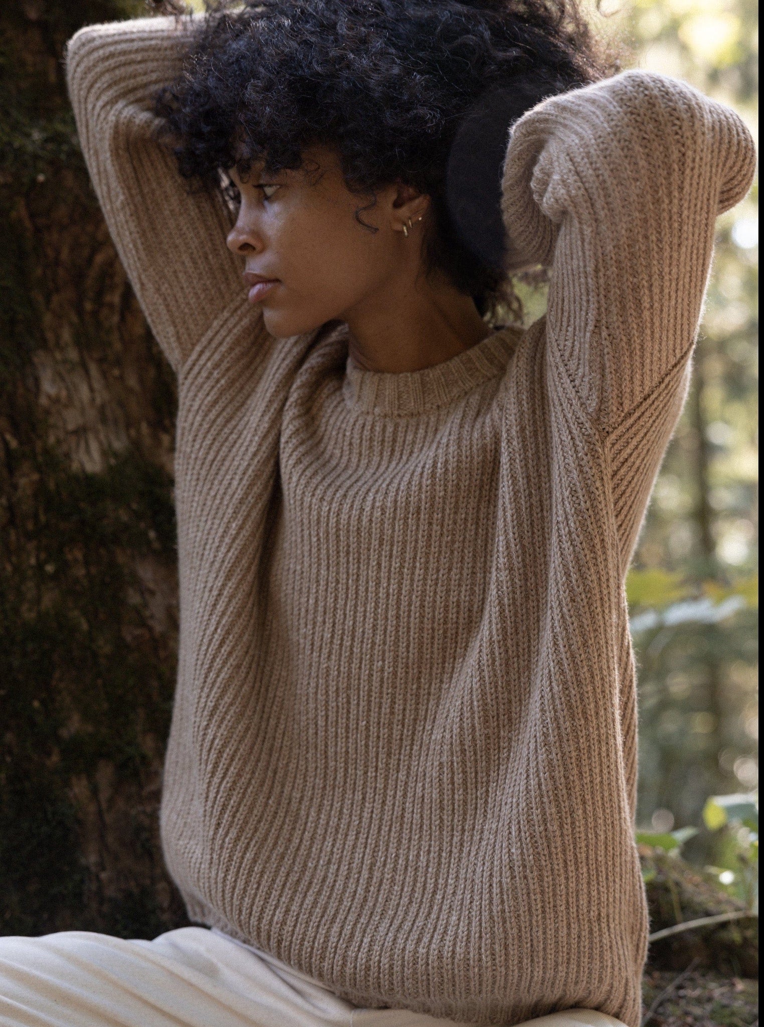 A woman in a Field Sweater - Caramel leaning against a tree in the woods.