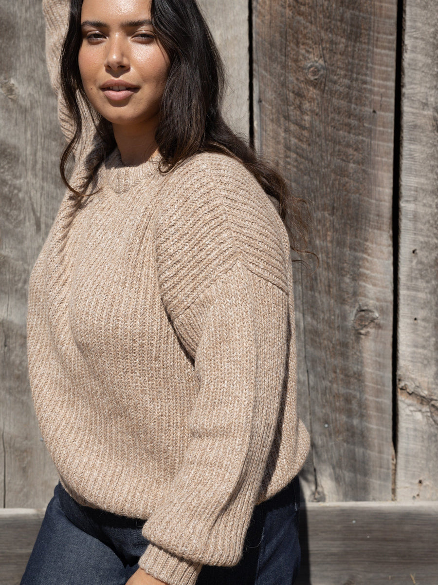 A woman wearing a Field Sweater - Caramel and jeans in front of a wooden fence.