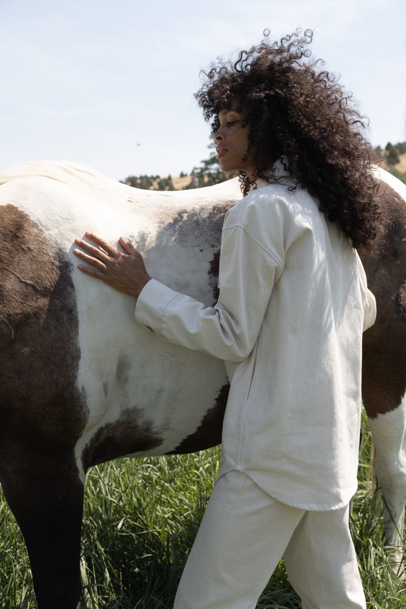 A woman in an upcycled denim Roan Shirt Jacket - Bone is seen petting a horse in a field.