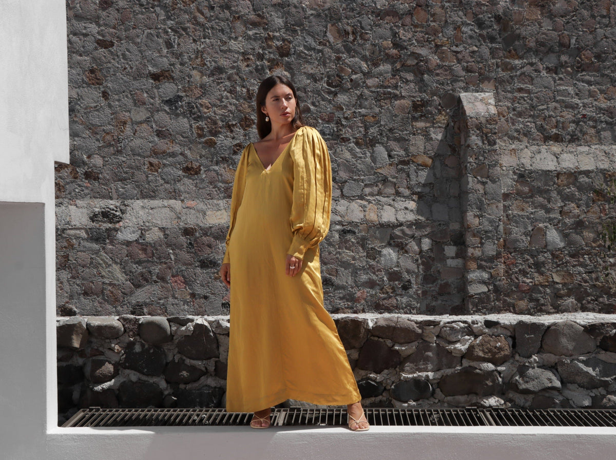A woman in a Lupita Maxi Dress - Citrine standing on a stone wall.