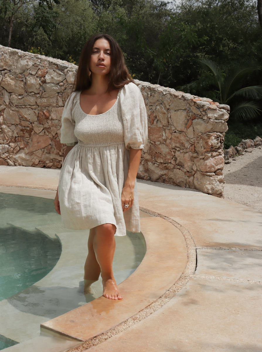 A woman in a white Carmen Mini Dress - Natural - Sample stands next to a pool under the Summer Sun.