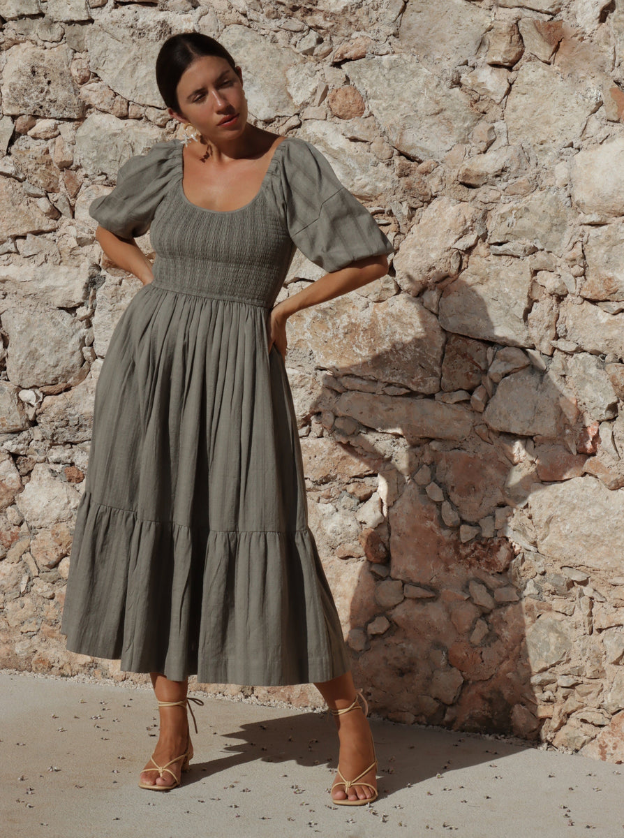 A woman wearing the Frida Midi Dress - Azure Broderie standing next to a stone wall.