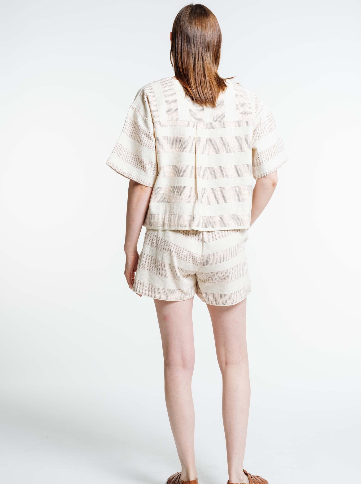 The back view of a woman wearing a Baker Top - Terracotta Ticking Stripe and shorts.